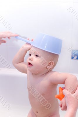 Baby in bath with toys put ladle on head like a hat