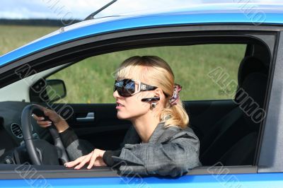 young blond woman in a blue car in sun-glasses with hands free bluetooth