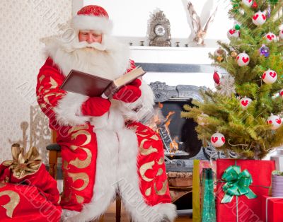 Santa sitting at the Christmas tree, near fireplace and reading.