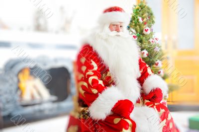 Santa sitting at the Christmas tree, near fireplace and looking at camera. 