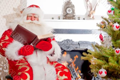 Santa sitting at the Christmas tree, near fireplace and reading 