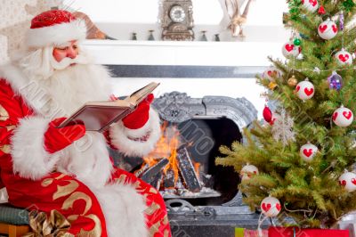 Santa sitting at the Christmas tree, near fireplace and reading 