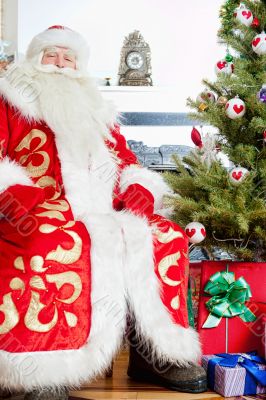 Santa sitting at the Christmas tree, fireplace and looking at ca