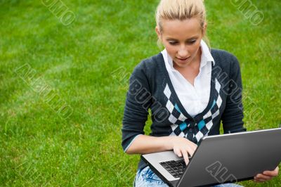 Young woman with laptop sitting on green grass and looking to th