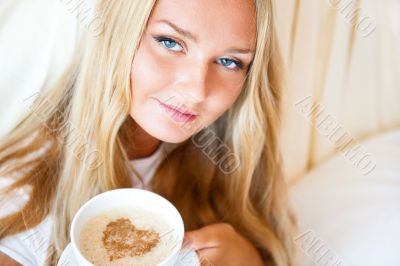 Smiling woman drinking a coffee lying on a bed at home or hotel.