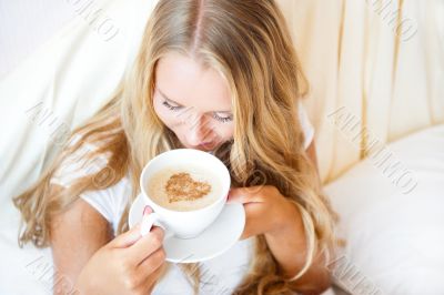 Smiling woman drinking a coffee lying on a bed at home or hotel.