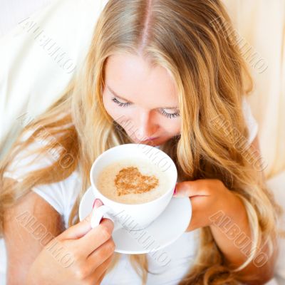 Smiling woman drinking a coffee lying on a bed at home or hotel.