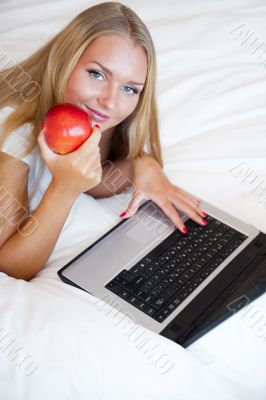 Smiling woman using a laptop while lying on her bed and eating r