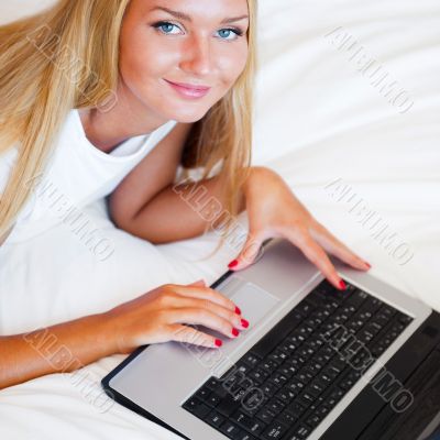 Smiling woman using a laptop while lying on her bed and eating r