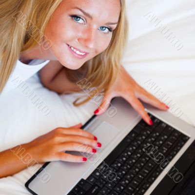 Smiling woman using a laptop while lying on her bed and eating r