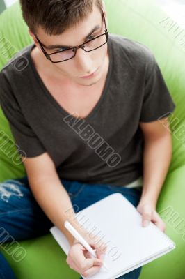 Young adult man writing in his copybook while sitting on a large