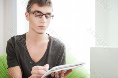 Young adult man writing in his copybook while sitting on a large