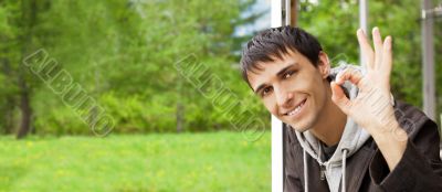 Young brunette man gives the OK sign, standing near big window a