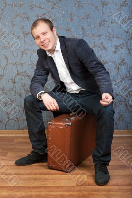 A young man sits on an old brown suitcase in the interior