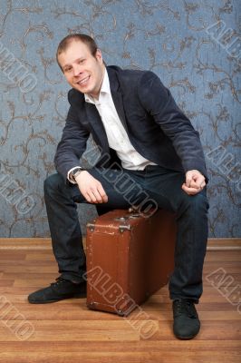 A young man sits on an old brown suitcase in the interior A young man sits on an old brown suitcase in the interior