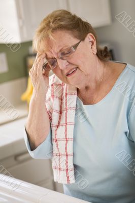 Senior Adult Woman At Kitchen Sink With Head Ache