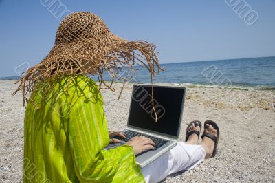 Woman in hat sitting on beach working on laptop
