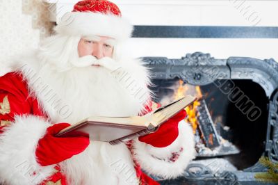 Santa sitting at the Christmas tree, near fireplace and reading.