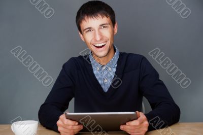 Closeup of a young smiling business man sitting at his office agagainst gray wall and using his modern tablet pc to do his work.