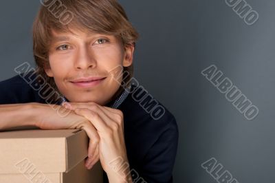 Portrait of young man leaning on box against grey wall. He is st