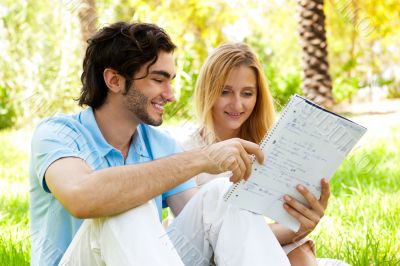 Happy couple of students with a notebook sitting on grass at cam