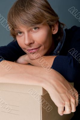 Portrait of young man leaning on box against grey wall. He is st
