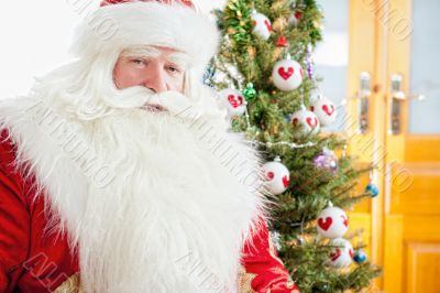 Santa sitting at the Christmas tree, fireplace and looking at ca