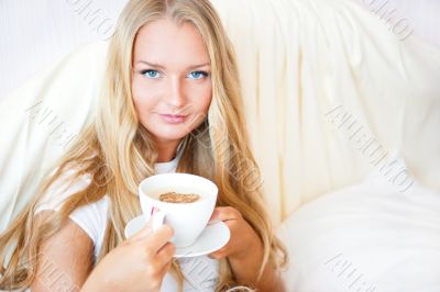 Smiling woman drinking a coffee lying on a bed at home or hotel.