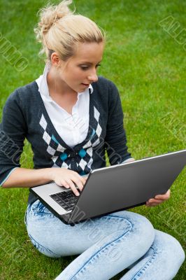 Young woman with laptop sitting on green grass and looking to th