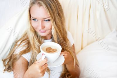 Smiling woman drinking a coffee lying on a bed at home or hotel.