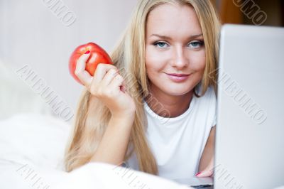 Smiling woman using a laptop while lying on her bed and eating r