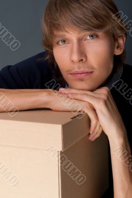 Portrait of young man leaning on box against grey wall. He is st