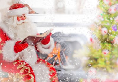 Santa sitting at the Christmas tree, near fireplace and reading 