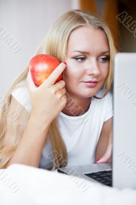 Smiling woman using a laptop while lying on her bed and eating r