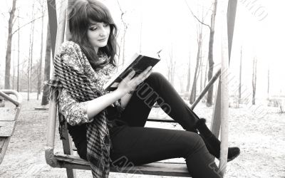 A beautiful woman is sitting in a park in the spring with a scarf on the shoulders of the trees on the swings and thoughtfully r