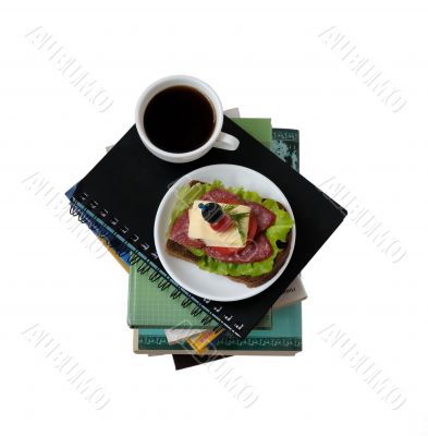 A stack of books and notebooks with a cup of coffee and a sandwi