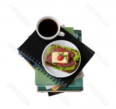 A stack of books and notebooks with a cup of coffee and a sandwi