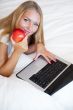 Smiling woman using a laptop while lying on her bed and eating r