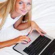 Smiling woman using a laptop while lying on her bed and eating r