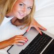 Smiling woman using a laptop while lying on her bed and eating r