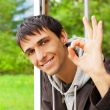 Young brunette man gives the OK sign, standing near big window a
