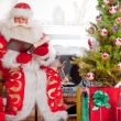 Santa sitting at the Christmas tree, near fireplace and reading 