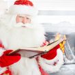 Santa sitting at the Christmas tree, near fireplace and reading 