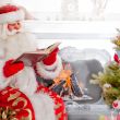Santa sitting at the Christmas tree, near fireplace and reading 