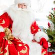Santa sitting at the Christmas tree, fireplace and looking at ca