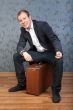 A young man sits on an old brown suitcase in the interior